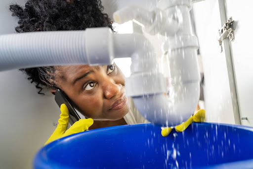 A woman looks frantically at a water leak. She is wearing gloves and calling a plumber.